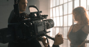 man filming woman with camera, windows in the background